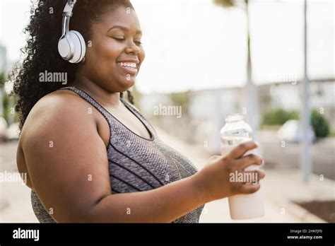 Curvy Woman Drinking While Doing Jogging Routine Outdoor At City Park Focus On Headphones