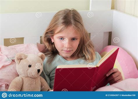 little blonde girl readingbook girl lying on white bed and reading book with her toy bear