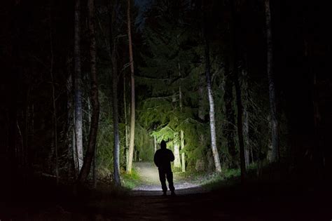 Man Standing In Dark Forest At Night With Flashlight And Hoodie On Head