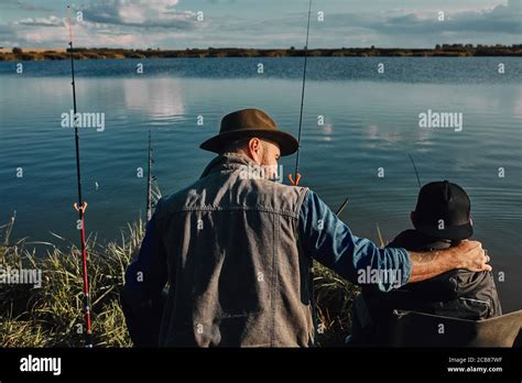 Backs Of Caucasian Father And Son Sit Near Lake On Fishing Chairs They