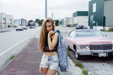 Sensual Blonde Girl Standing On The Sidewalk Looking At The Camera Near To A Classic Pink Car