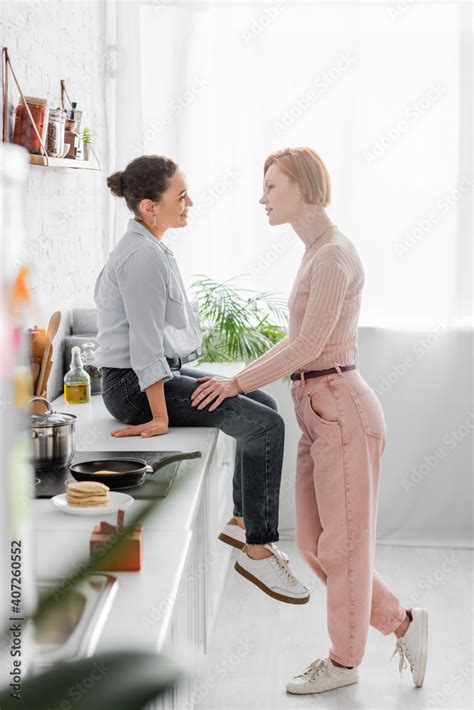 Side View Of Redhead Lesbian Woman Talking To African American Girlfriend Sitting On Kitchen