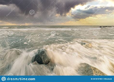 Waves Wash The Rocks On The Sandy Beach With A Cloudscape In The