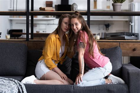 dos lesbianas sonrientes sentadas en el sofá y mirando a cámara en el salón foto de archivo