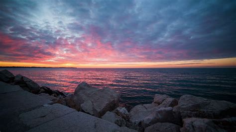 Rocks On Beach With Pink Sunset On Sea Wide Shot Of Rocks And Pier In Front And Pink Sea And