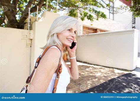 Older Woman Walking And Talking With Mobile Phone On Sidewalk Stock