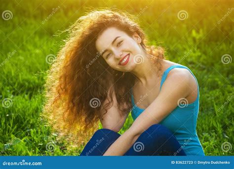 Attractive Young Girl With Curly Hair Sitting On The Green Grass On The Lawn And Smiling At The