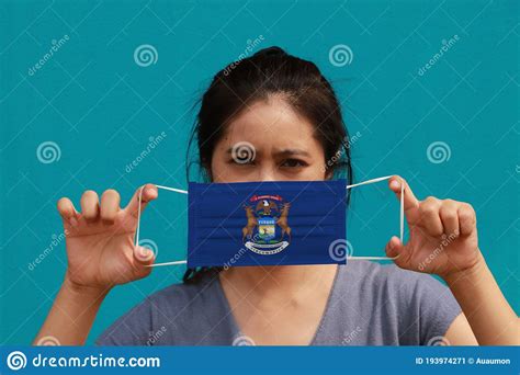 a woman and hygienic mask with michigan flag pattern in her hand and raises it to cover her face