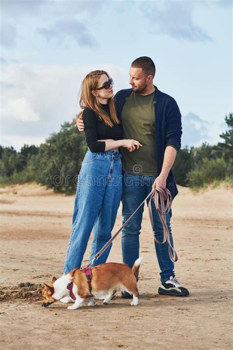 Young Happy Couple And Dog Stand On Beach Against Pines And Sand