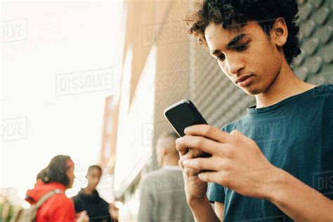 Teenage Boy Using Mobile Phone While Friends Standing In Background On