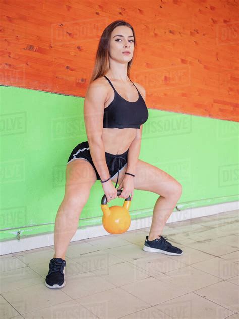fit blond female in sportswear holding heavy kettlebell and looking at camera while standing