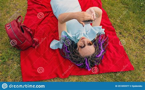 Young Woman Lying On A Blanket Blowing Uses A Smartphone In The Park