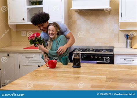 Happy Biracial Young Husband Giving Roses To Beautiful Wife Having Coffee At Table In Kitchen