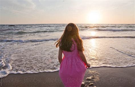 Girl In Pink Dress Standing At Oceans Edge Facing Sunset By Dina Marie
