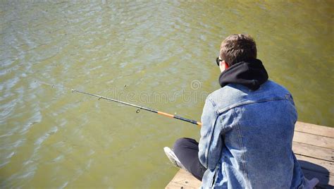 Young Fisherman In Denim Jacket Sitting On Wooden Pier Fishing In The