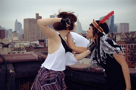 Two Women Standing On Top Of A Roof Looking At Each Other With City In