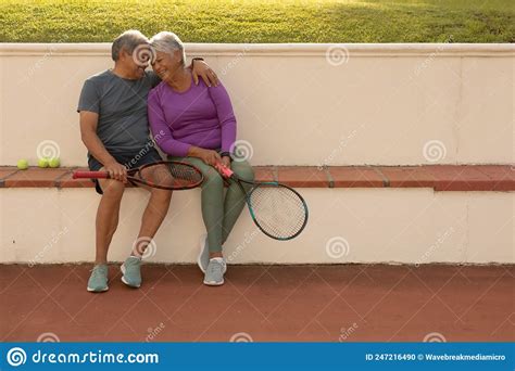 Happy Romantic Biracial Senior Couple Holding Tennis Rackets While Sitting On Bench At Tennis