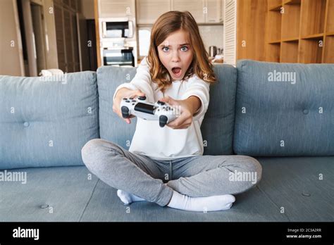 photo of scared caucasian girl playing video game with joystick while sitting on couch at home