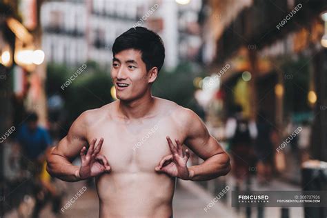 Playful Asian Shirtless Guy Gesturing And Covering Nipples While Standing On Urban Street In