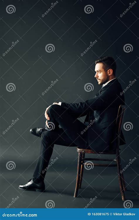 Confident Handsome Man In Black Suit Sitting On The Chair On Dark