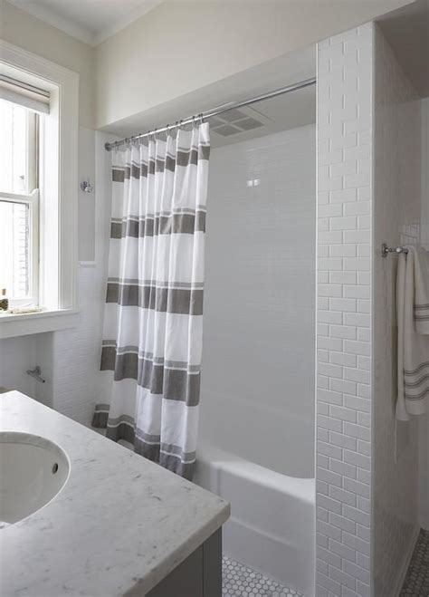 A luxurious white marble bathroom with a large window with curtains, a tub clad with marble and a shower space. A gray stripe shower curtain hangs over marble hex floor ...