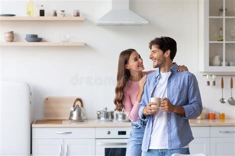 Cheerful Millennial European Wife Hugs Husband With Cup Of Hot Drink In