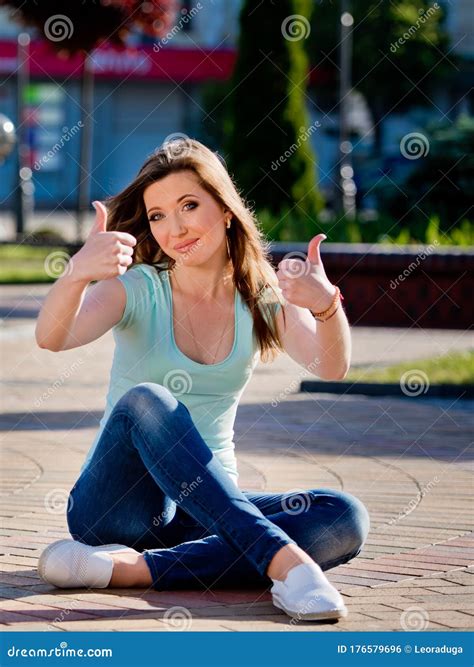 A Young And Pretty Girl In Jeans Sits On A Sidewalk Tile In The Middle