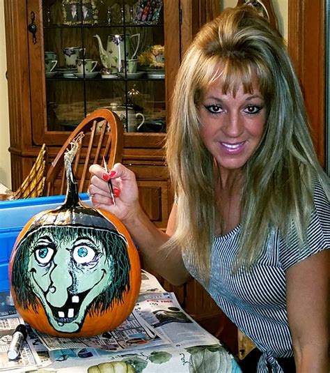 A Woman Holding Up A Painted Pumpkin With An Evil Face On It In Front Of A Dining Room Table