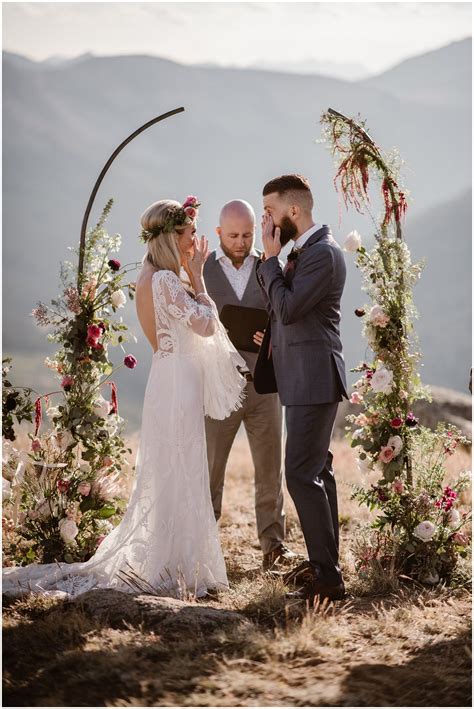 A Bride And A Groom Both Stand In Front Of A Gorgeous Flowered Arch As