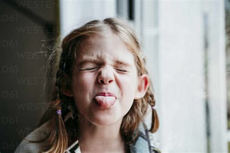 Blond Girl Sticking Tongue Out While Standing By Window At Home Stock Photo