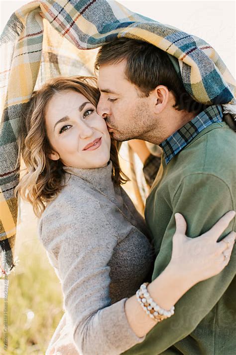 A Husband Kissing His Wife On The Cheek Under A Plaid Blanket By