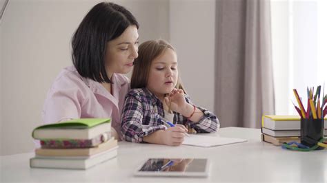 Side View Of Brunette Caucasian Mother Helping Daughter To Write Down Pretty Schoolgirl Doing