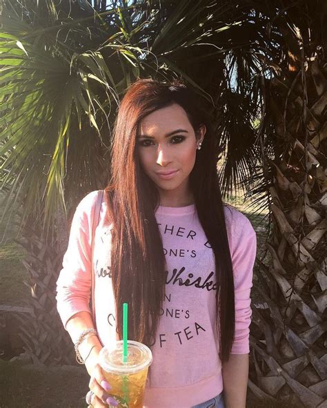 A Woman With Long Hair Holding A Drink In Front Of Some Palm Trees And Looking At The Camera