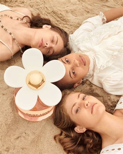 Two Women Laying On The Sand With A Perfume Bottle And Flower In Front Of Them