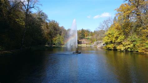 Fountain With Rainbow In Middle Of Lake Between Trees With Yellow