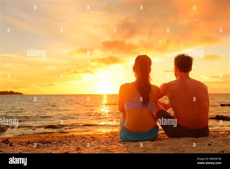 Happy Romantic Couple Enjoying Beautiful Sunset At The Beach Sitting In Sand Looking At Ocean