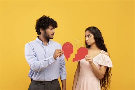Disappointed Indian Man And Woman Holding Two Pieces Of Broken Paper Heart Standing Over Yellow