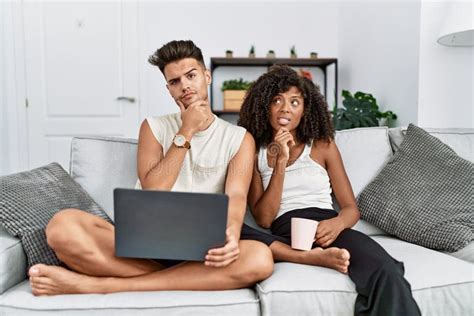 Young Interracial Couple Using Laptop At Home Sitting On The Sofa Thinking Worried About A