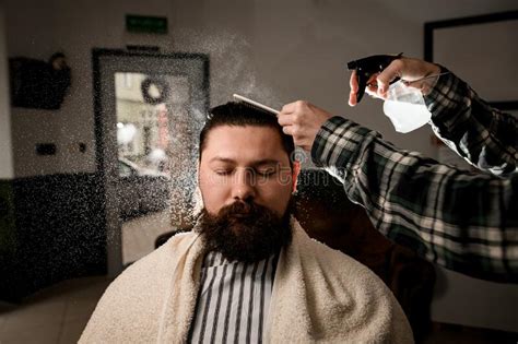 portrait of bearded man getting haircut and barber wets hair by spray and combs it barbershop