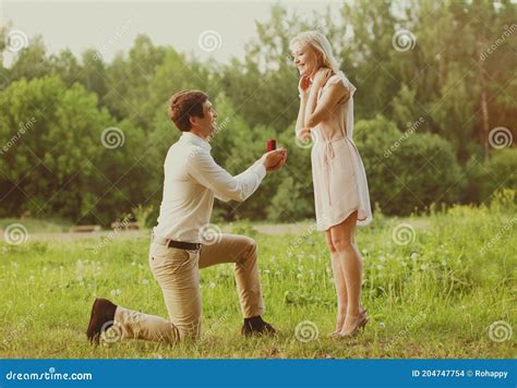 Happy Couple Man Kneeling Down And Proposing Ring To His Woman Outdoors On The Grass Wedding