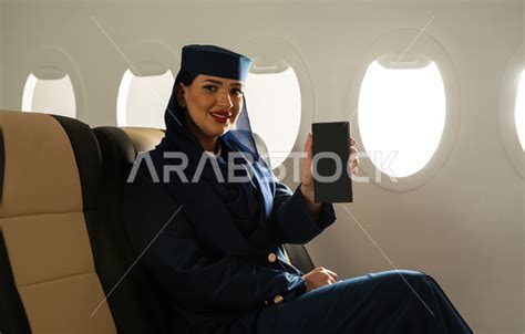 مضيفة طيران عربية سعودية خليجية تجلس في مقعد الطائرة وتمسك هاتف محمول بشاشة سوداء فارغة، عرض