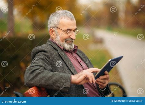 Grandpa Uses A Tablet Sitting In The Park On The Bench Stock Photo