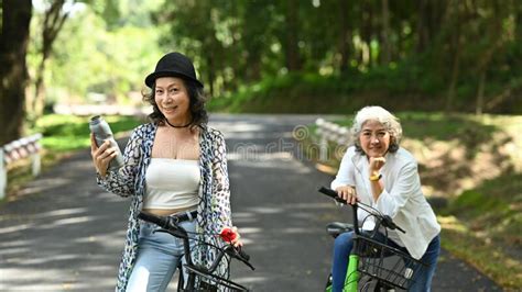 Two Smiling Middle Aged Women Riding Bicycles Through Public Park Together Retirement People