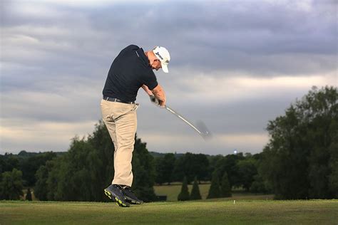 A Man Swinging A Golf Club On Top Of A Lush Green Field With Trees In