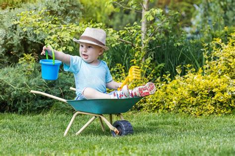 Cute Baby Boy Is Sitting On Green Wheelbarrow Holding A Blue Bucket