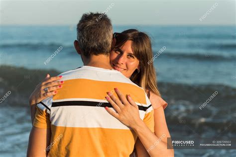 adult man hugging woman while standing on beach near waving sea and resting together — affection
