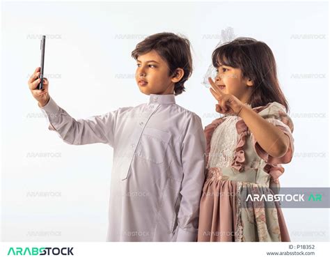 خلفية بيضاء لصبي وفتاه صغيران سعوديان مبتسمان ، يمسك الصبي الجوال بيده يقومان بإلتقاط صور