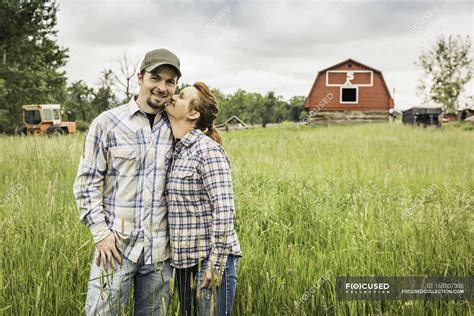 Couple On Farm In Tall Grass Looking At Camera Smiling Kiss On Cheek