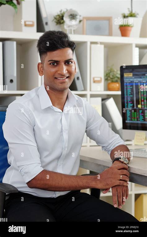 Portrait Of Handsome Young Indian Trader In White Shirt Sitting At Office Table With Stock