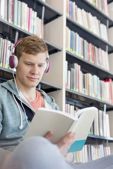 Student Reading Book While Listening Music Against Bookshelf At Library